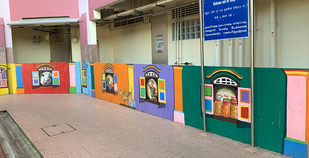 Low wall with mural of colourful windows side by side, each with an object from Little India peeking through