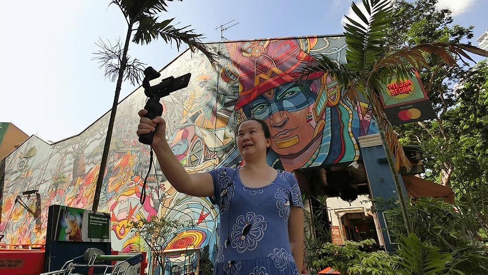 Me holding a gimbal and mobile phone in front of a colourful mural in Kampong Gelam