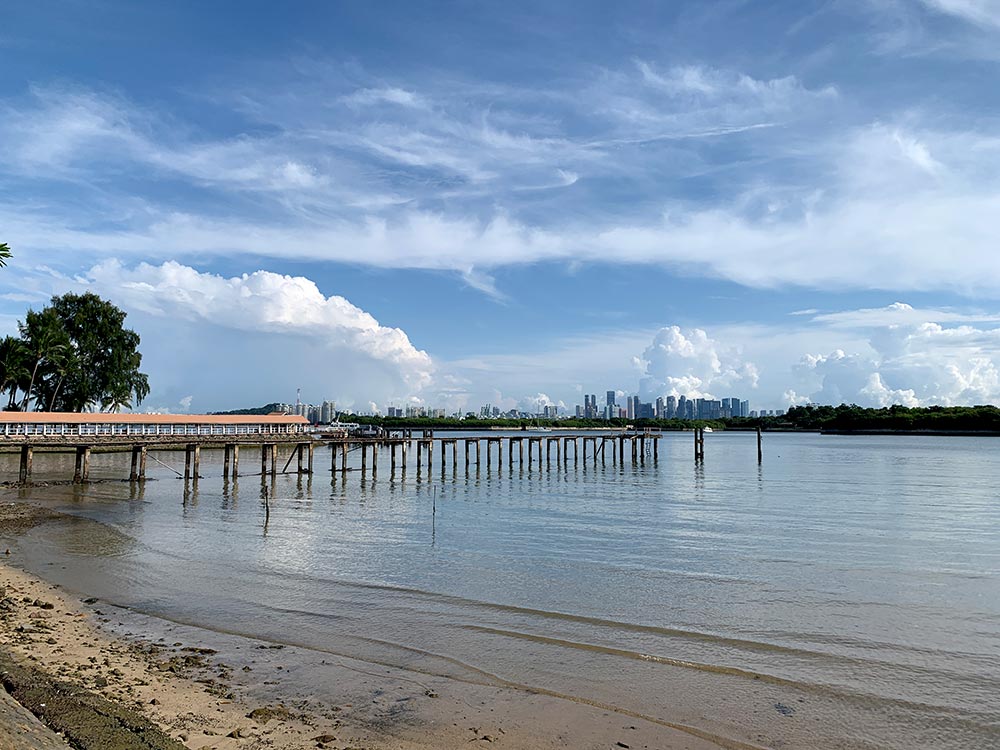 The old wooden posts of the old jetty extend out to sea parallel to the current jetty. Singapore's skyline can be seen in the background on this fine day.